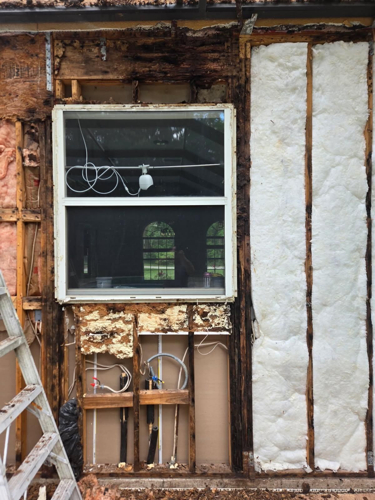 Exterior wall under renovation, exposing framing, insulation, and a window; visible electrical and plumbing.