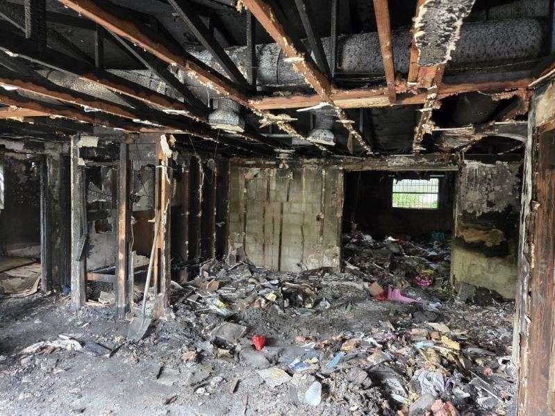 Interior of a fire-damaged room: charred walls, exposed ceiling beams, debris scattered on the floor.