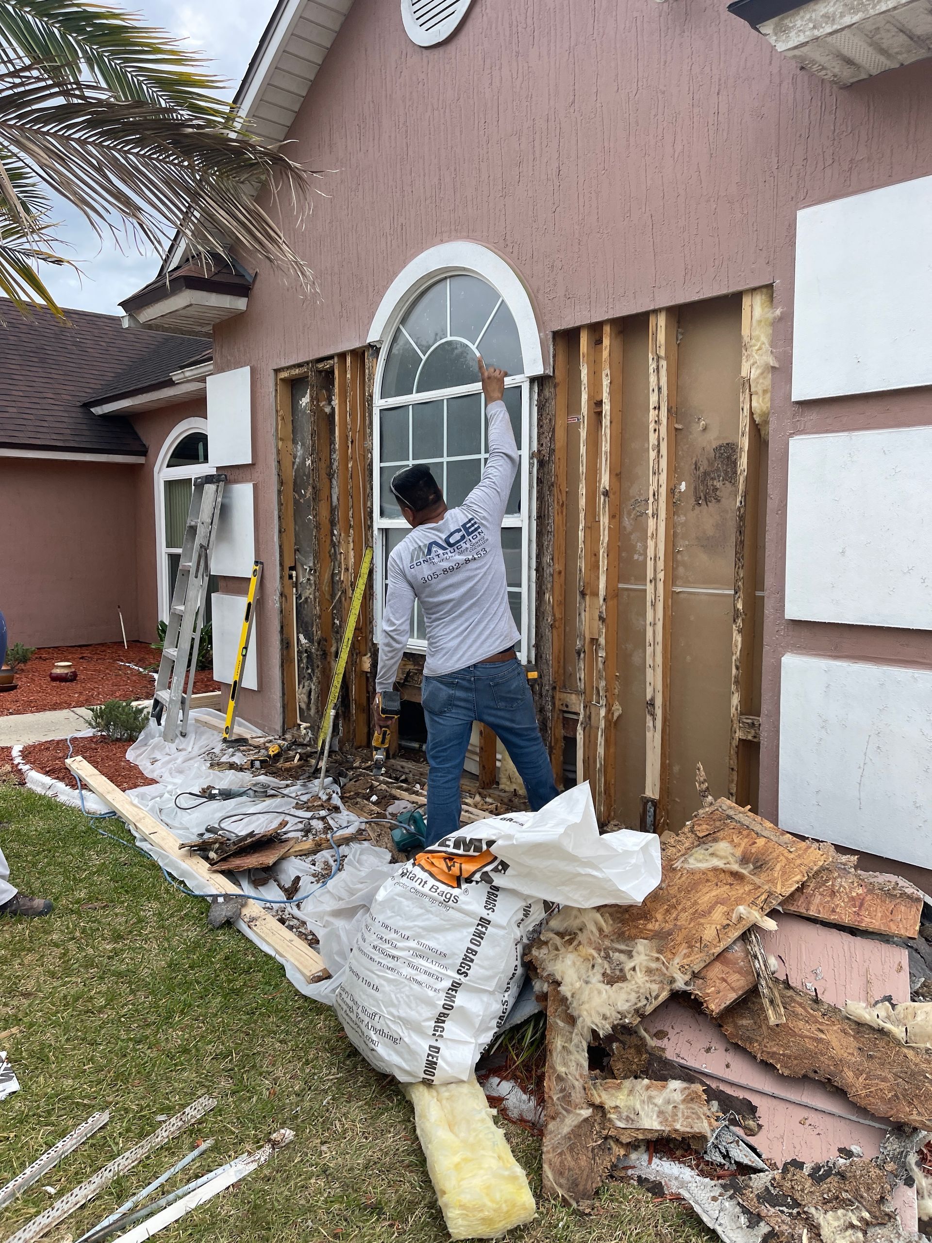 A worker stands next to a house with damaged siding. He's wearing a grey shirt and jeans, and is inspecting the exposed frame.