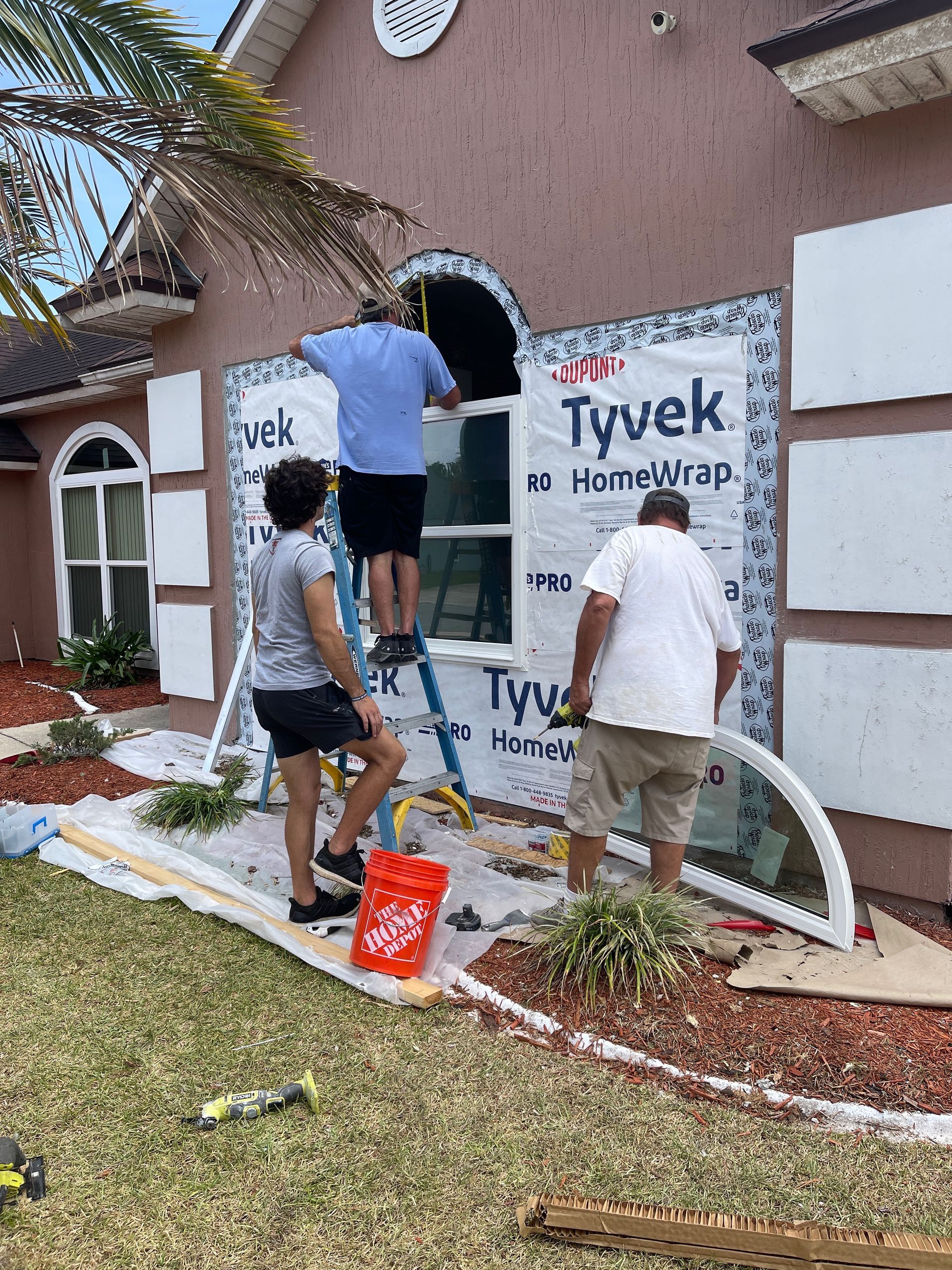Three people installing a window. One on a ladder, applying Tyvek. One holds the glass, the other watches. Outdoors, by a pink stucco house.