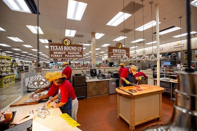 Inside a Buc-ee's store, employees in red shirts and hats prepare food behind counters. Overhead signs read