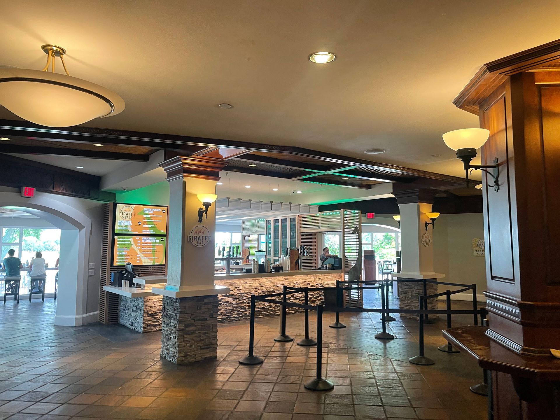An interior shot of a bar area. Features a bar with a stone facade, tables, and stanchions. The area has an arched entrance.