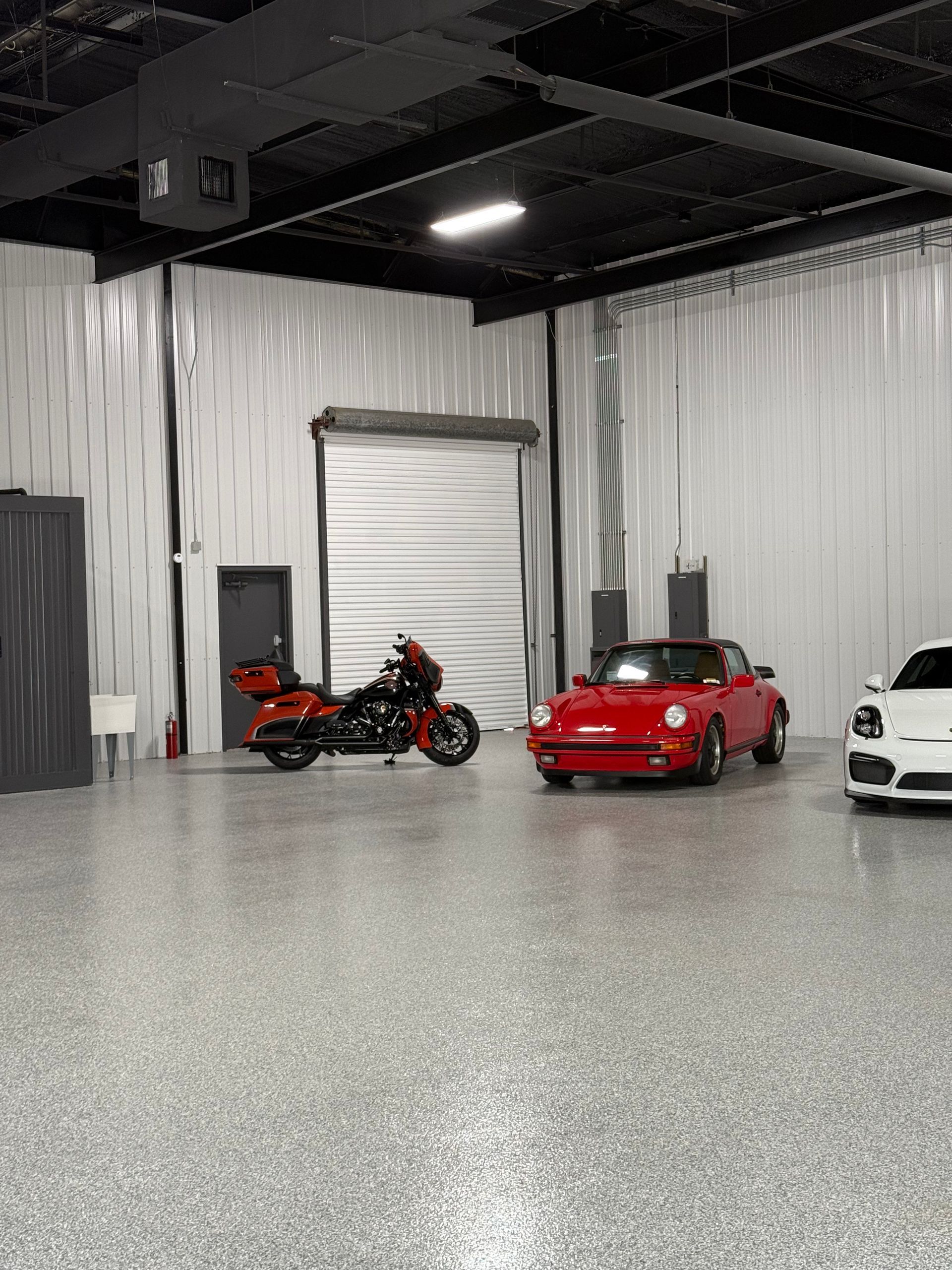 A spacious garage with a red Porsche, black motorcycle, and white car parked on a speckled floor, along with a roller door.