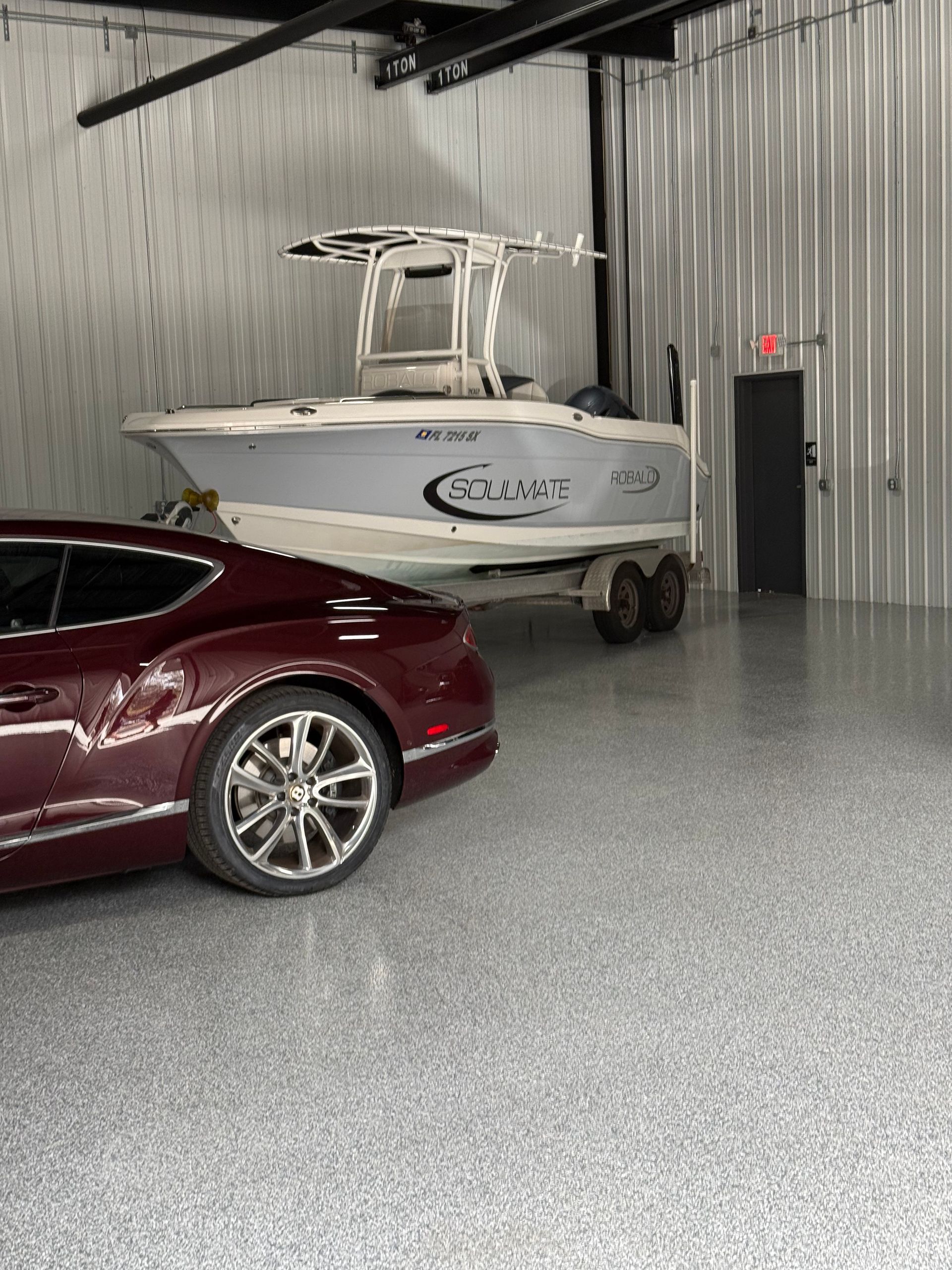 A luxury maroon car parked in a garage with a boat on a trailer. The garage has metallic walls and a shiny floor.