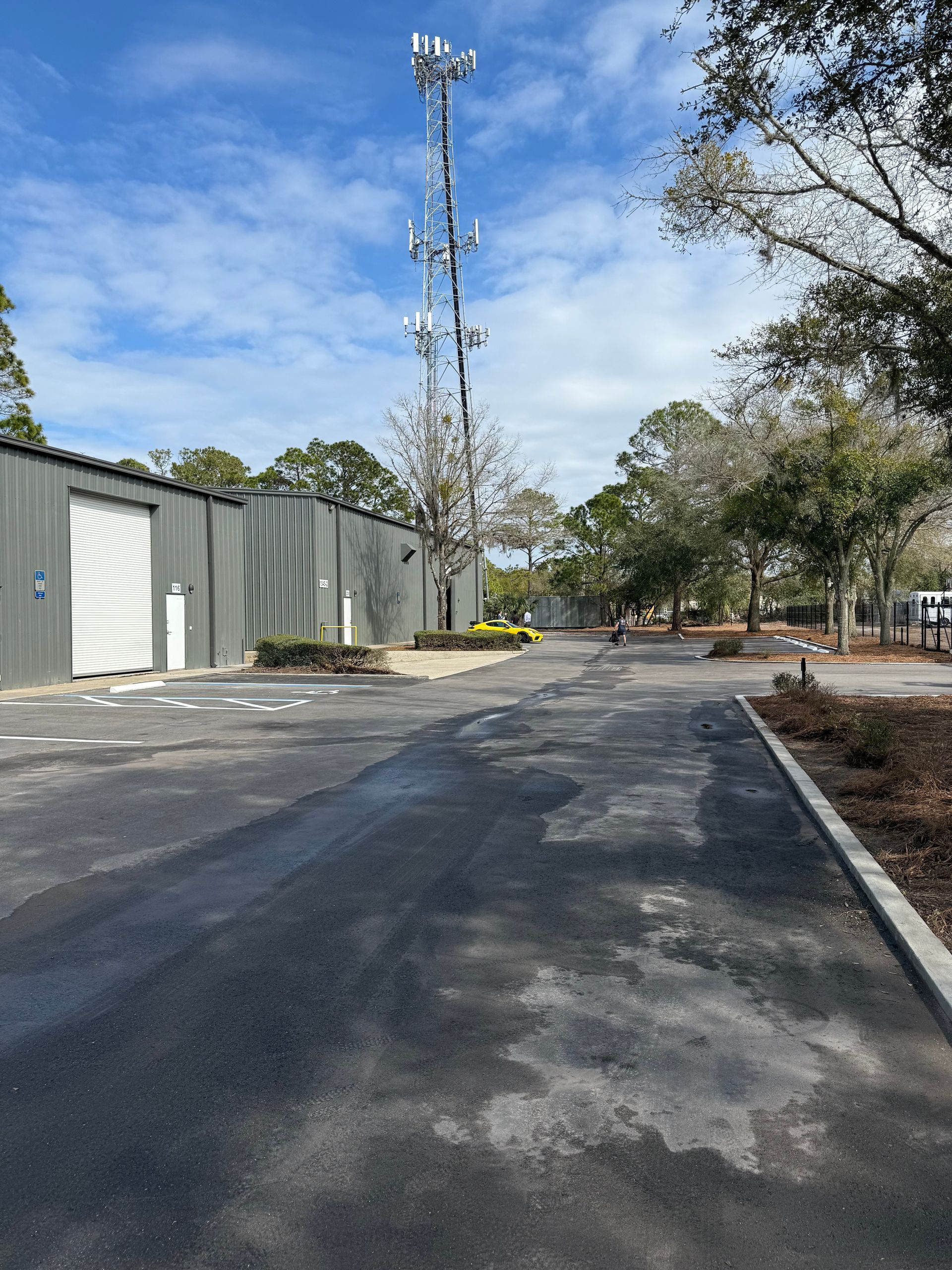 Dark asphalt driveway leads to a metal building and a cell tower against a partly cloudy sky. Trees line the right side.