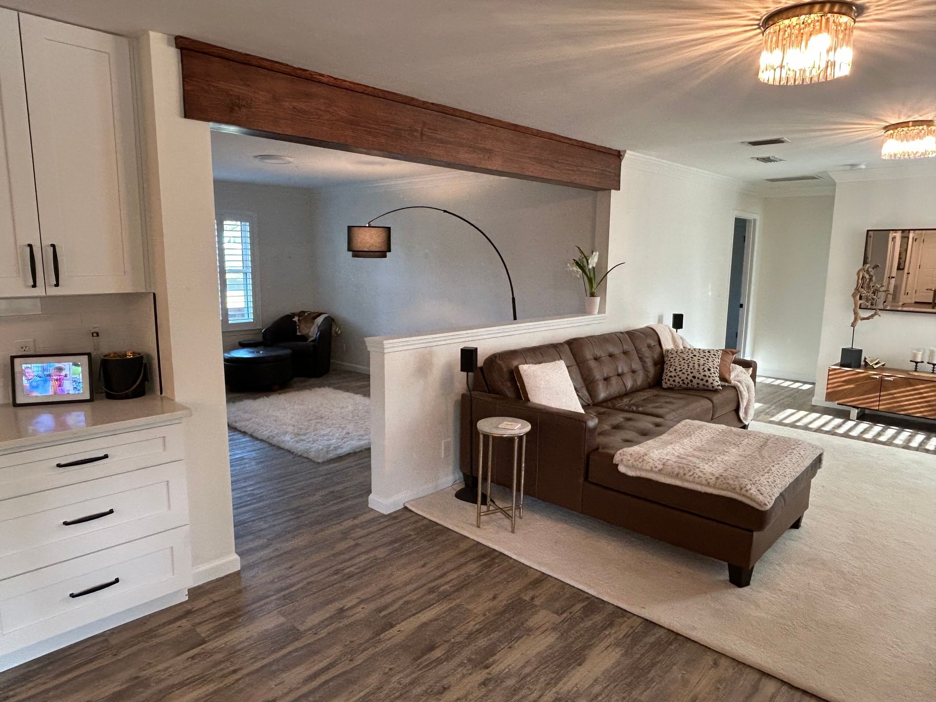 A modern living room with a brown leather chaise lounge, white walls, and wood-look flooring. A half-wall separates the seating area from a view of another room.