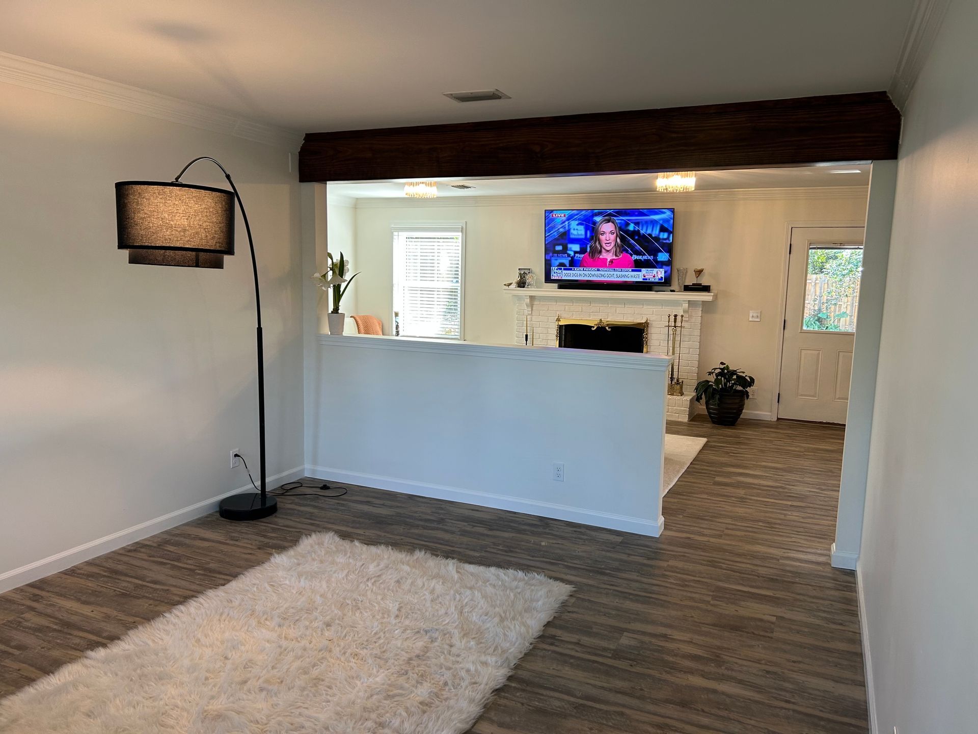 Living room with white walls, dark wood beam, and wood-look floor. A TV is mounted above a fireplace in the background.