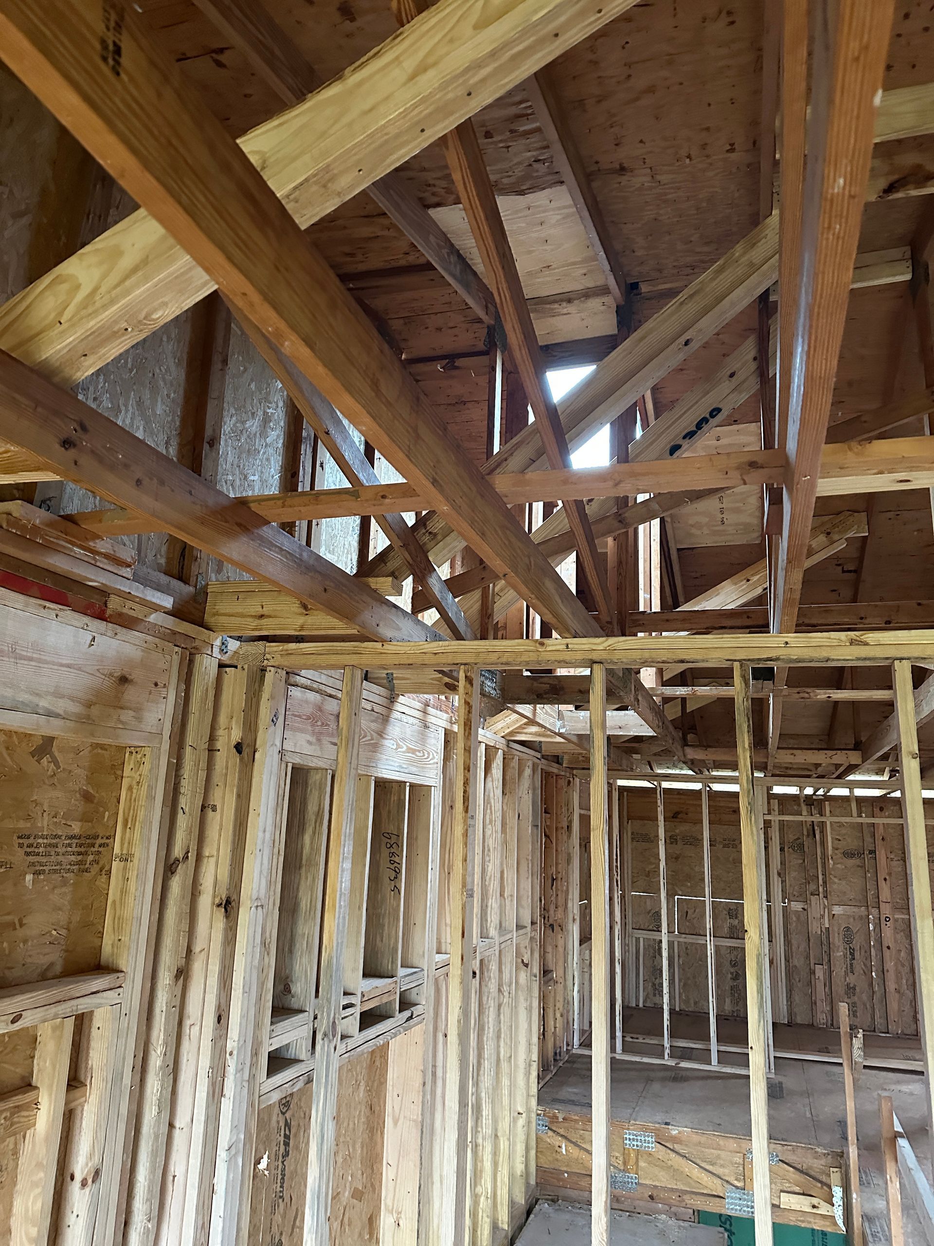 Interior view of a building under construction, showing exposed wooden framing for walls and roof. Daylight visible through the open roof structure.