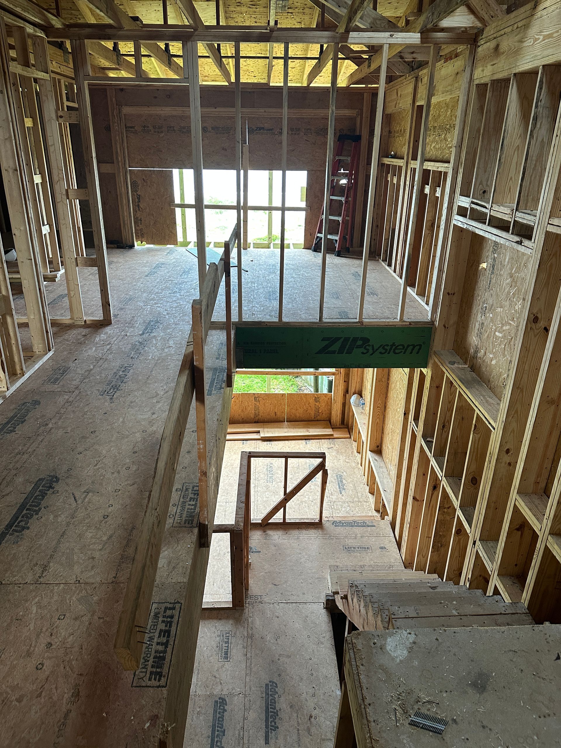 Interior view of a home under construction. Wooden framing and unfinished floors are visible. Sunlight streams through window openings in the distance.