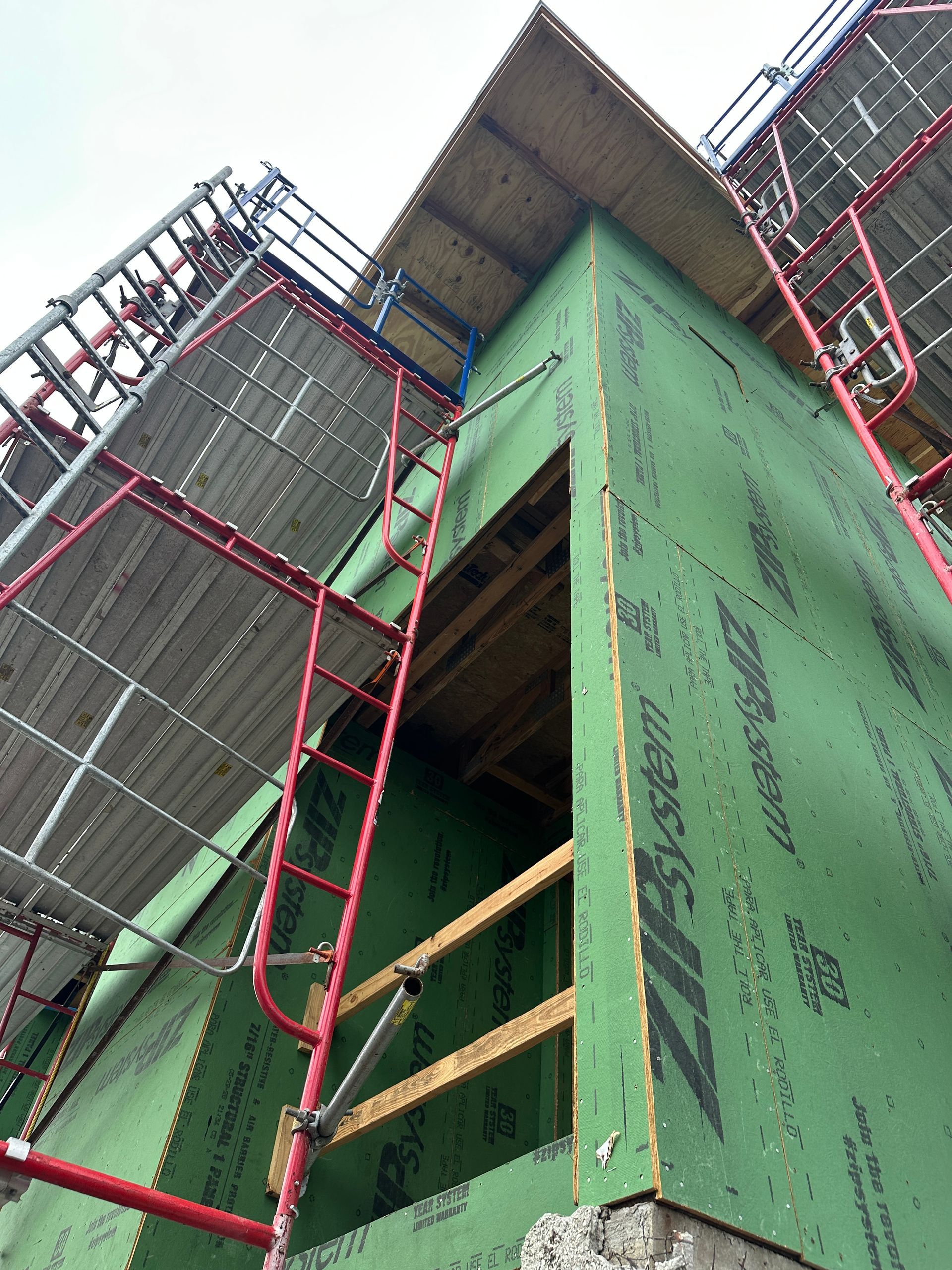 Construction site exterior, showing a building corner covered in green sheathing. Red scaffolding surrounds the building.