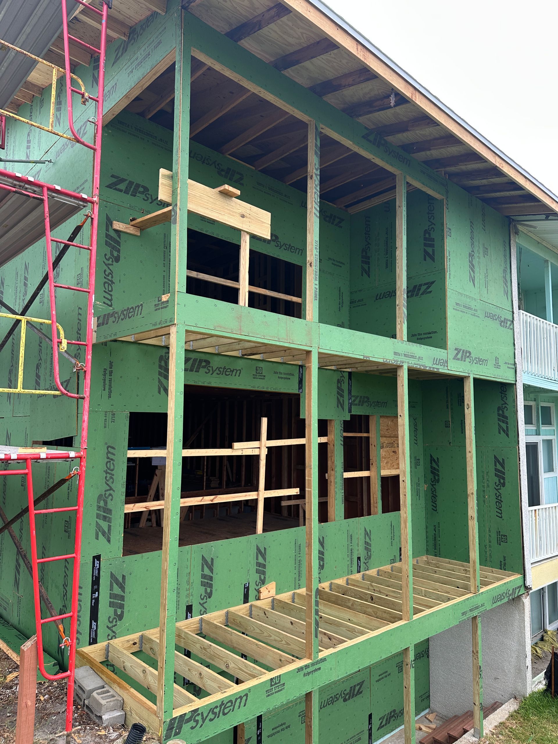 A building under construction with green siding and wood framing for an addition, including a deck and second-story balcony.