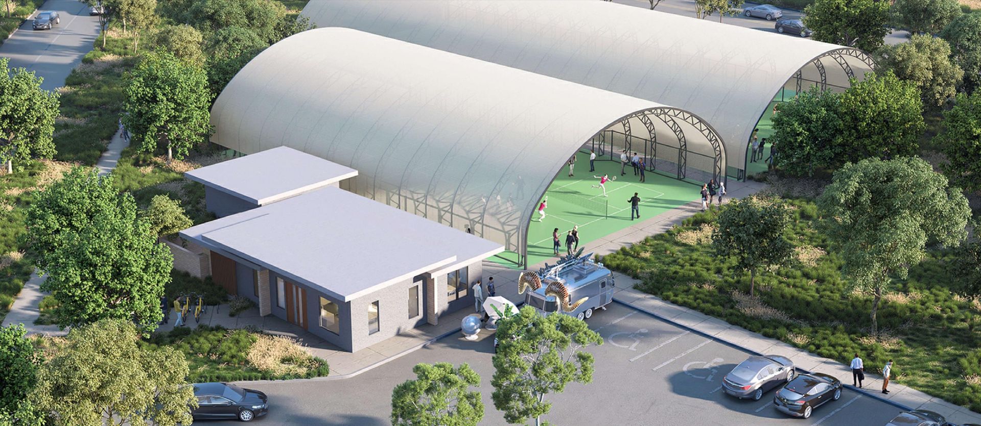 An aerial view of a modern building complex with two arched-roofed structures, a low-lying building, and a parking lot surrounded by trees.