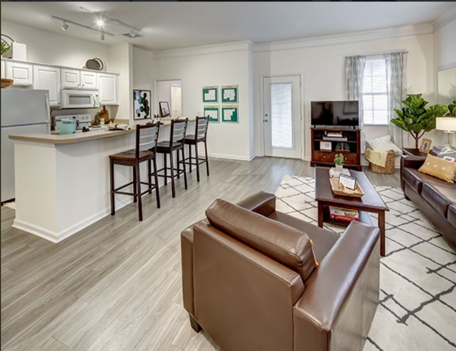 A furnished apartment with a kitchen, living area, and a brown leather armchair. Light wood-look floors and white walls.