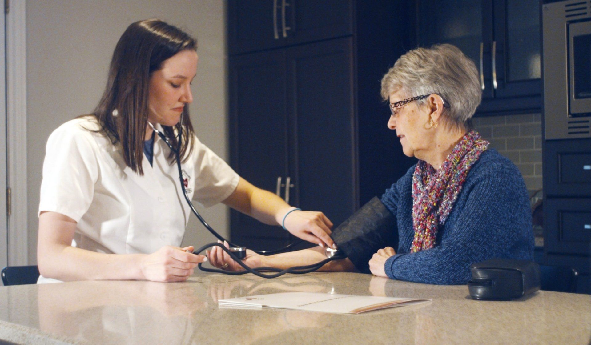 Nurse checking an elderly woman's blood pressure at a kitchen counter.