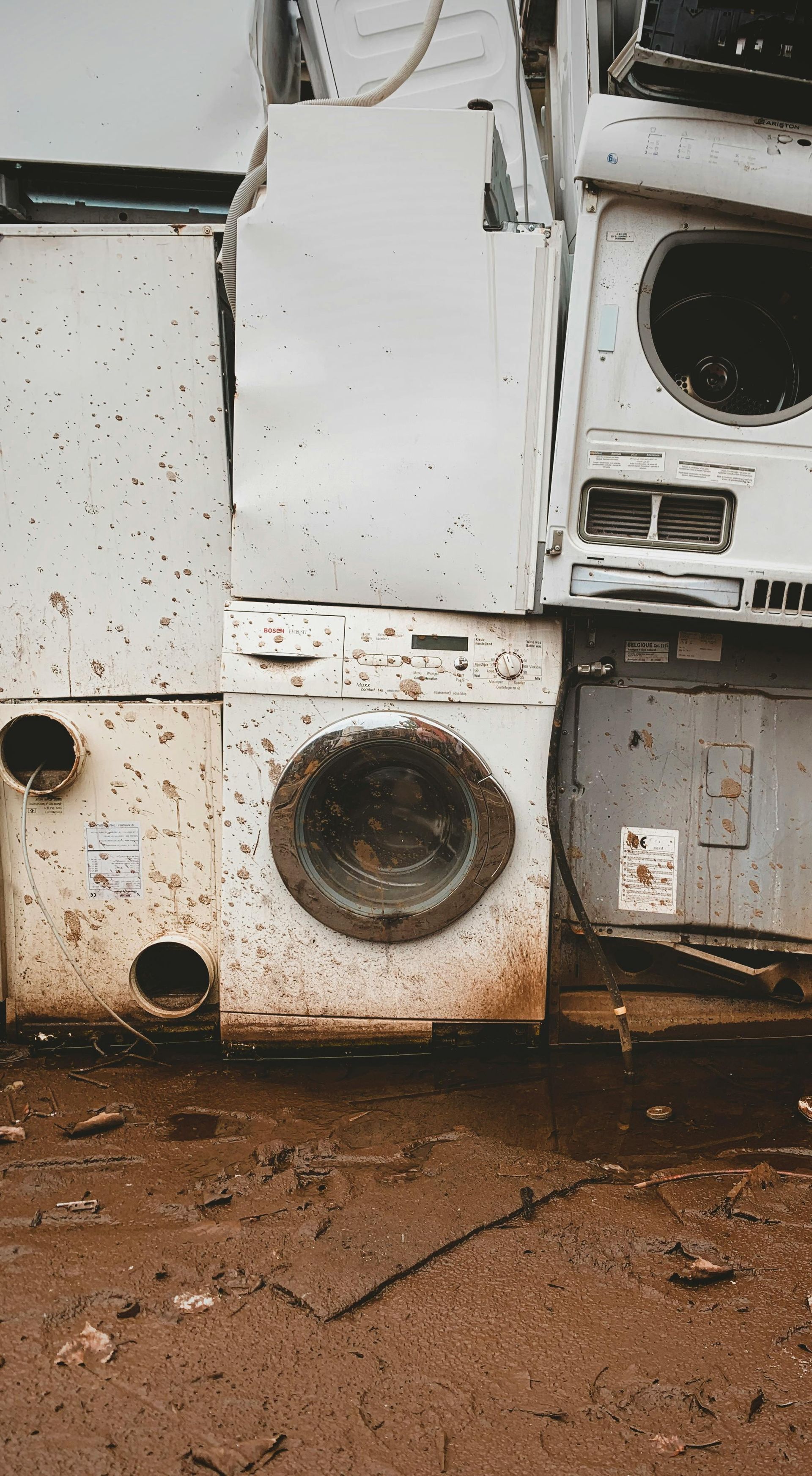 A pile of discarded, weathered white appliances, including washing machines, sits in a muddy, debris-filled outdoor area.