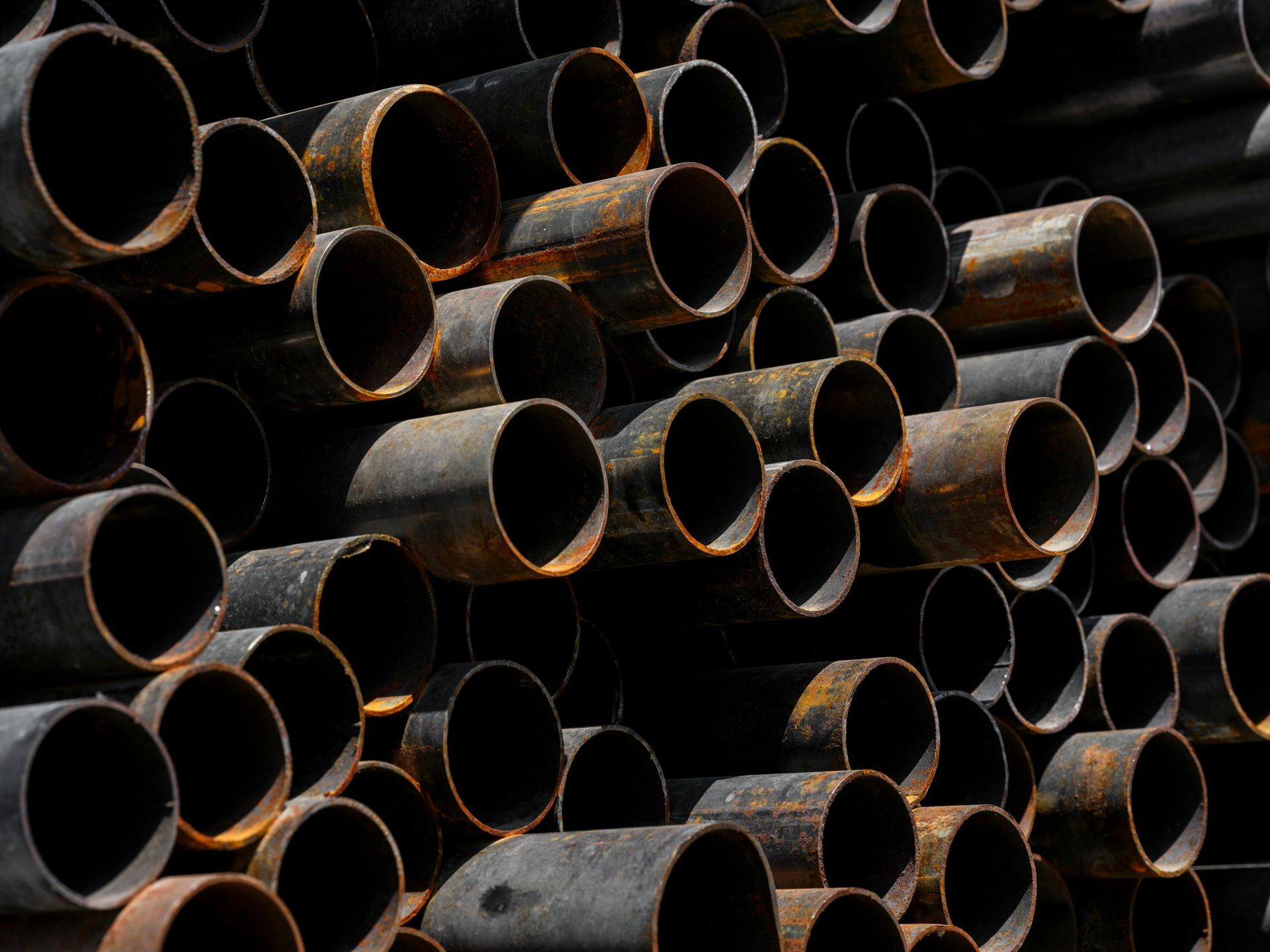 A large stack of dark, weathered cylindrical metal pipes piled together.