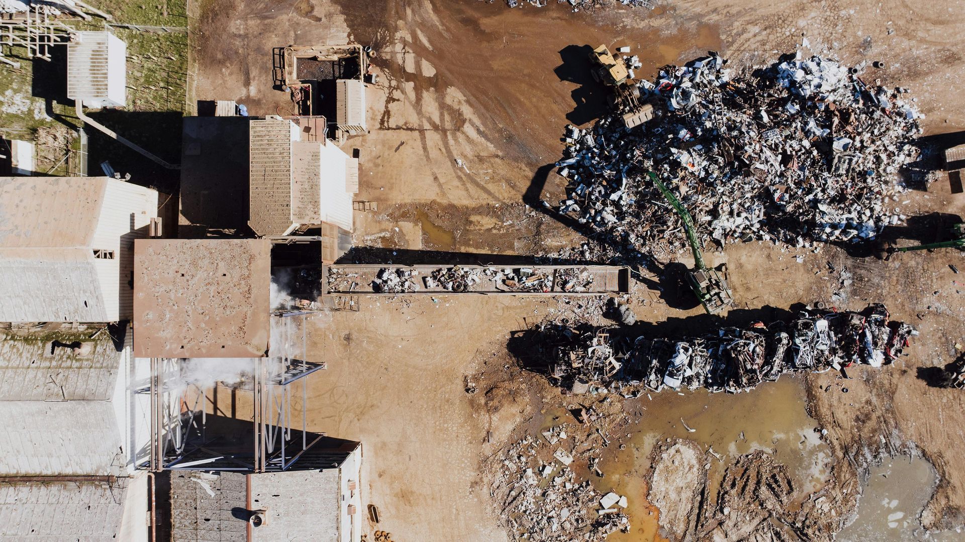 Aerial view of a scrapyard with piles of metal debris, a conveyor belt, and buildings on a dusty ground.