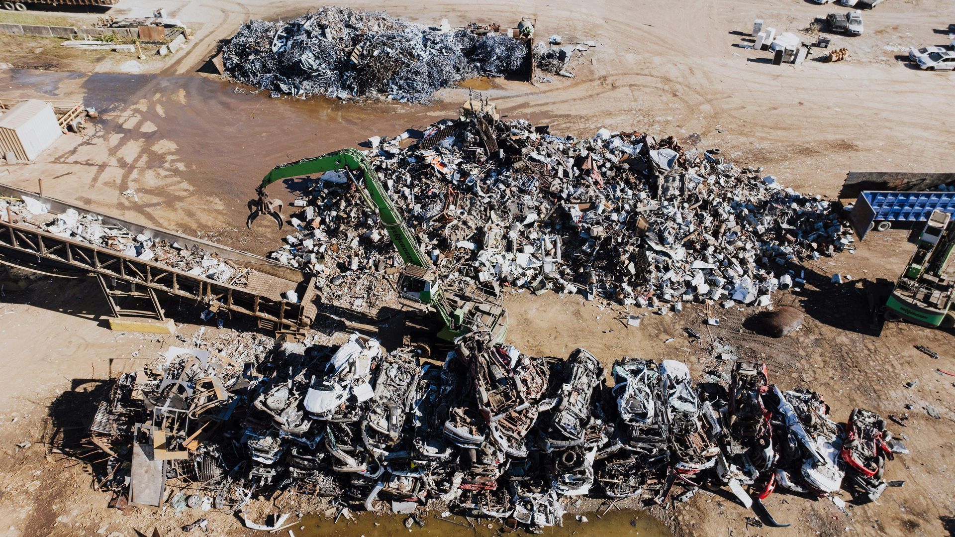 An aerial view shows a scrapyard where a green crane sorts piles of crushed metal and discarded vehicle parts.
