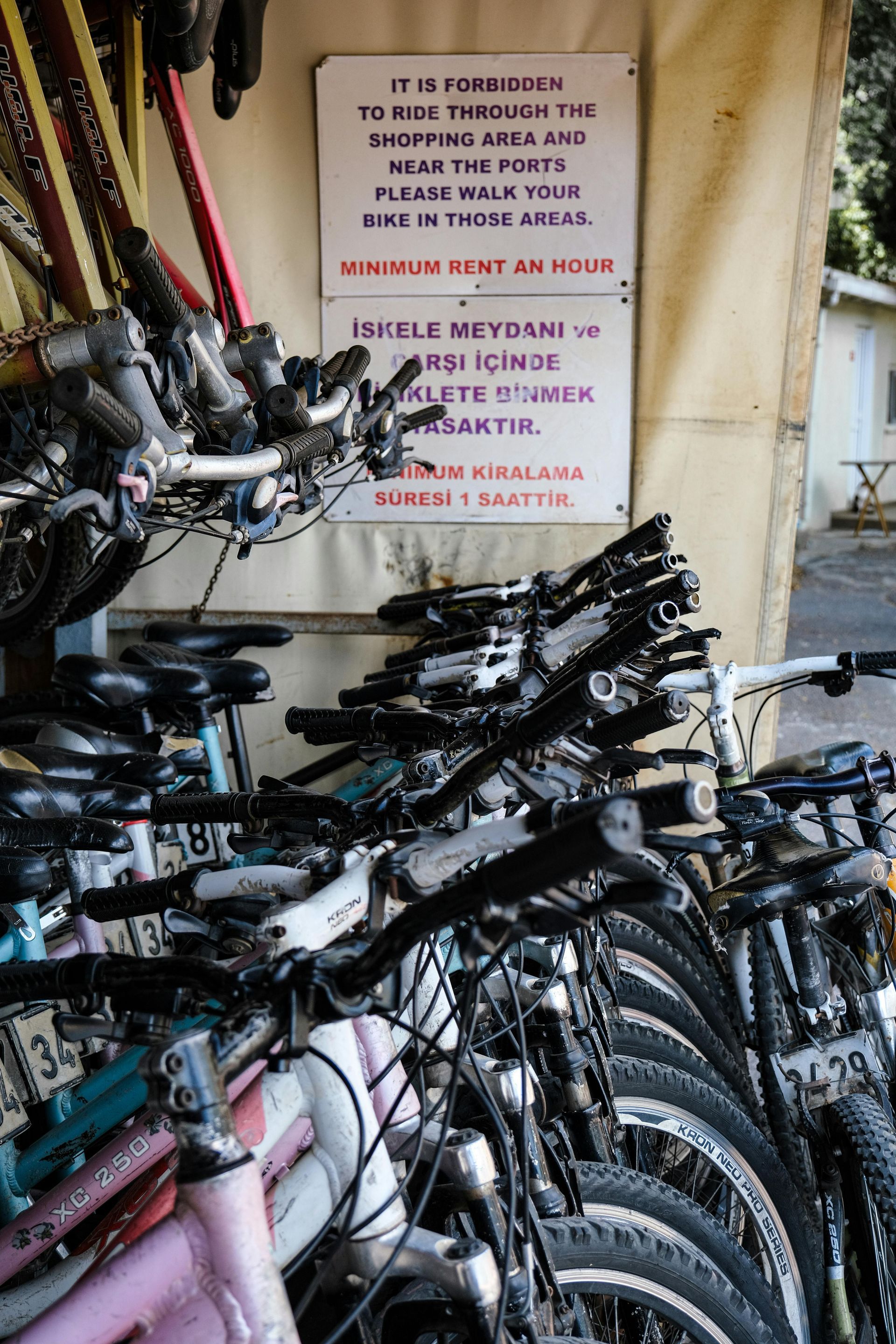 A sign in English and Turkish warns against riding bicycles in a shopping area, placed above a row of parked rental bikes.