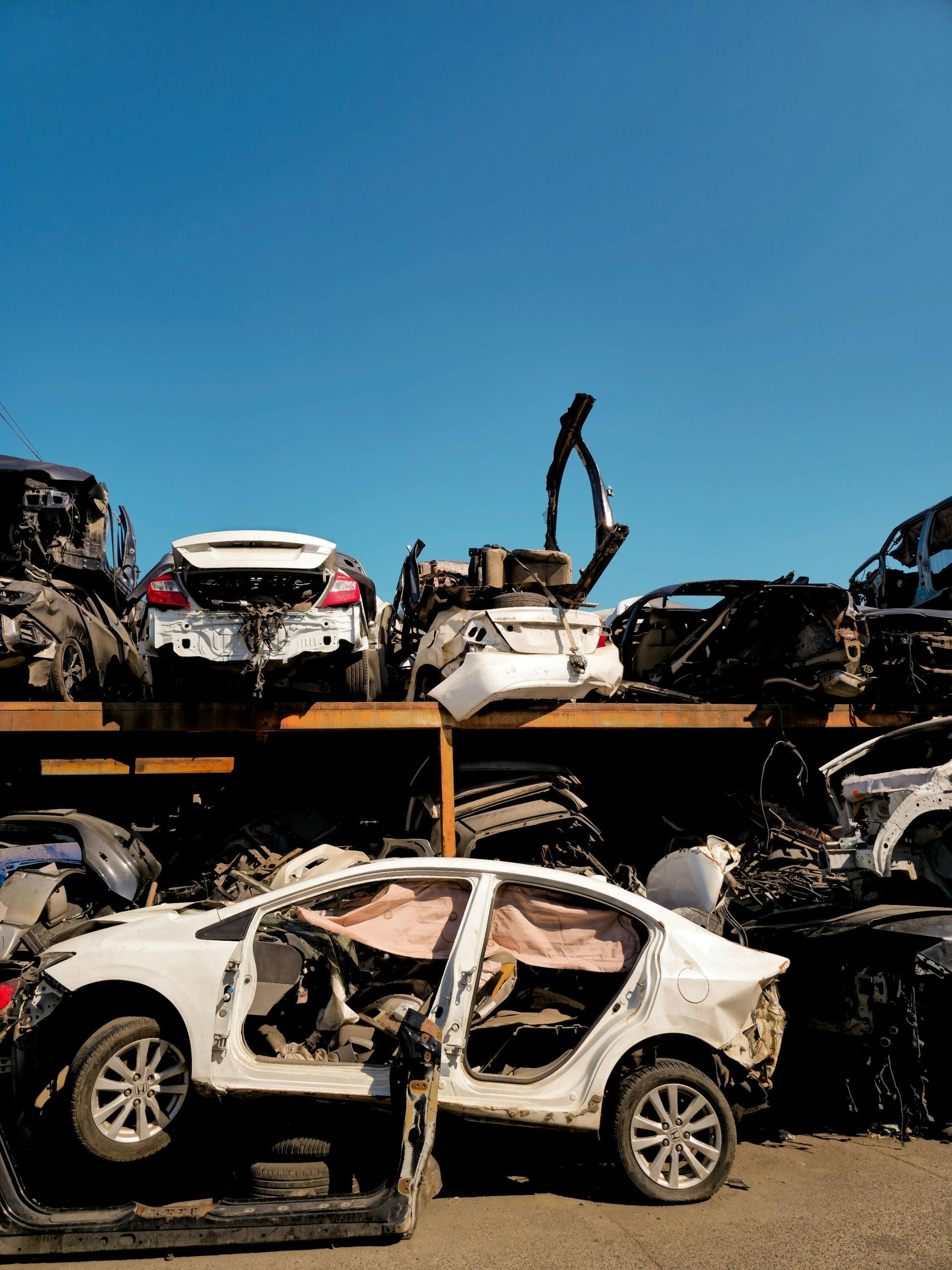 Stacked white cars at a vehicle salvage yard under a bright, clear blue sky.