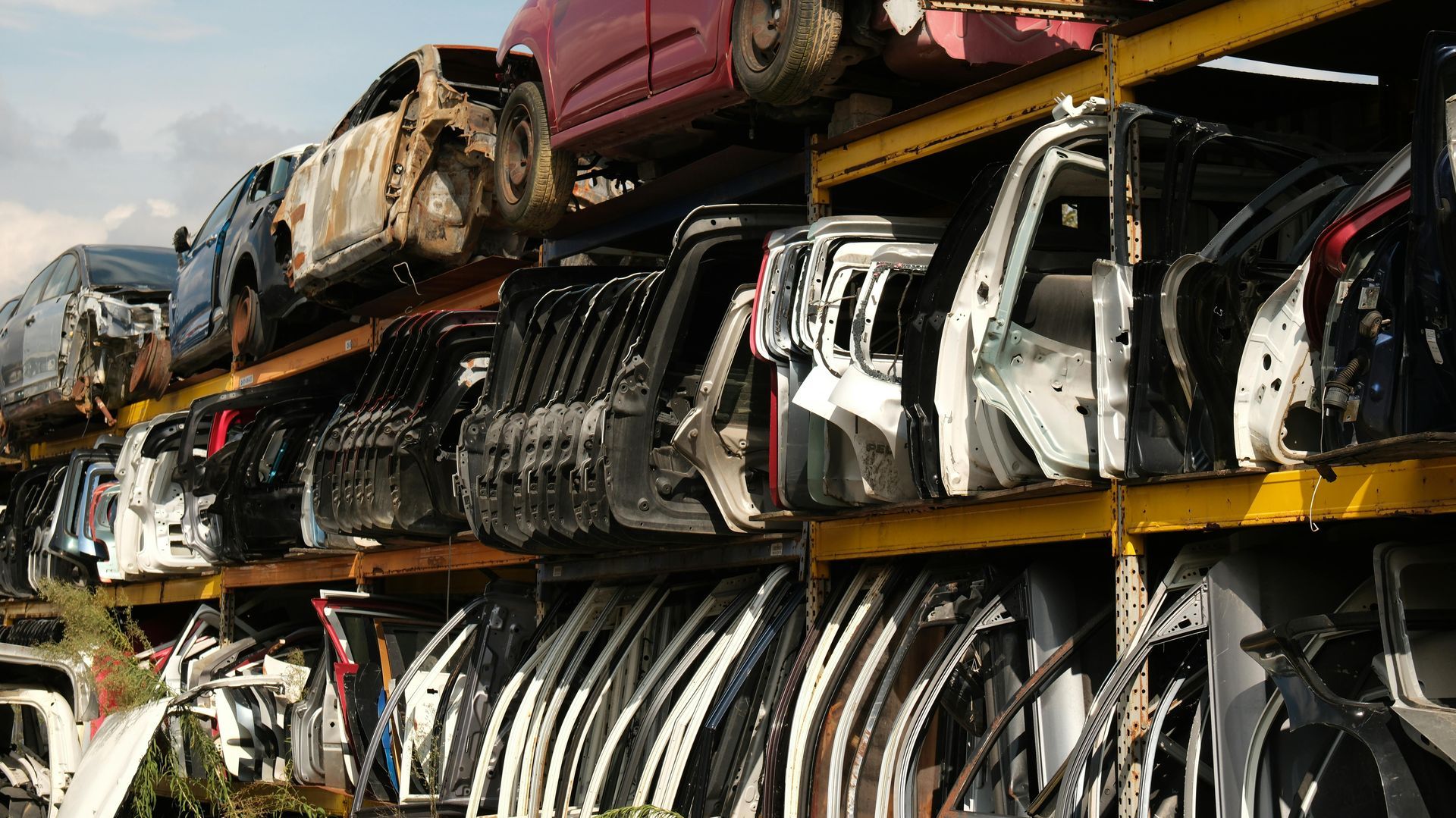 A metal shelving unit in an outdoor salvage yard, stacked with rows of salvaged car doors and vehicle parts.