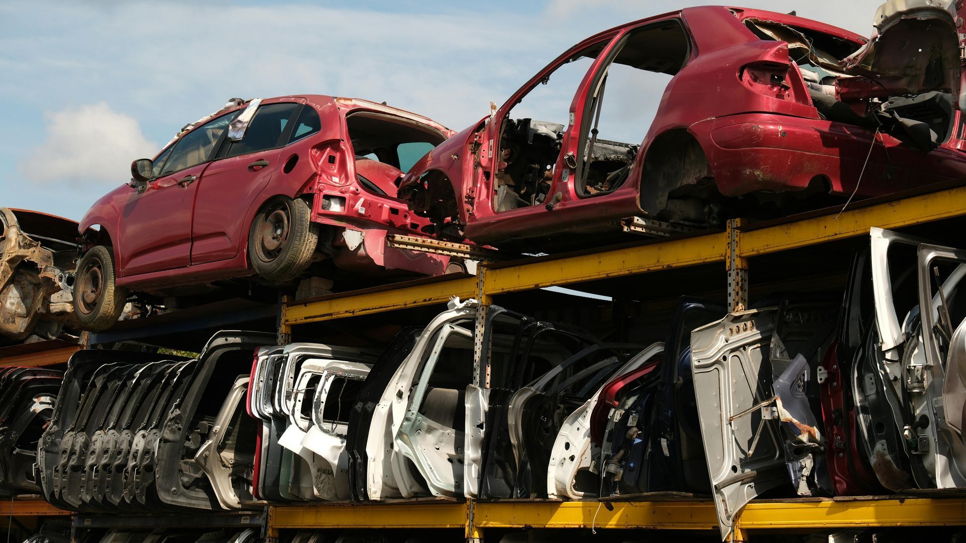 Red car shells and assorted salvaged vehicle parts stacked on industrial metal shelving in a scrapyard.
