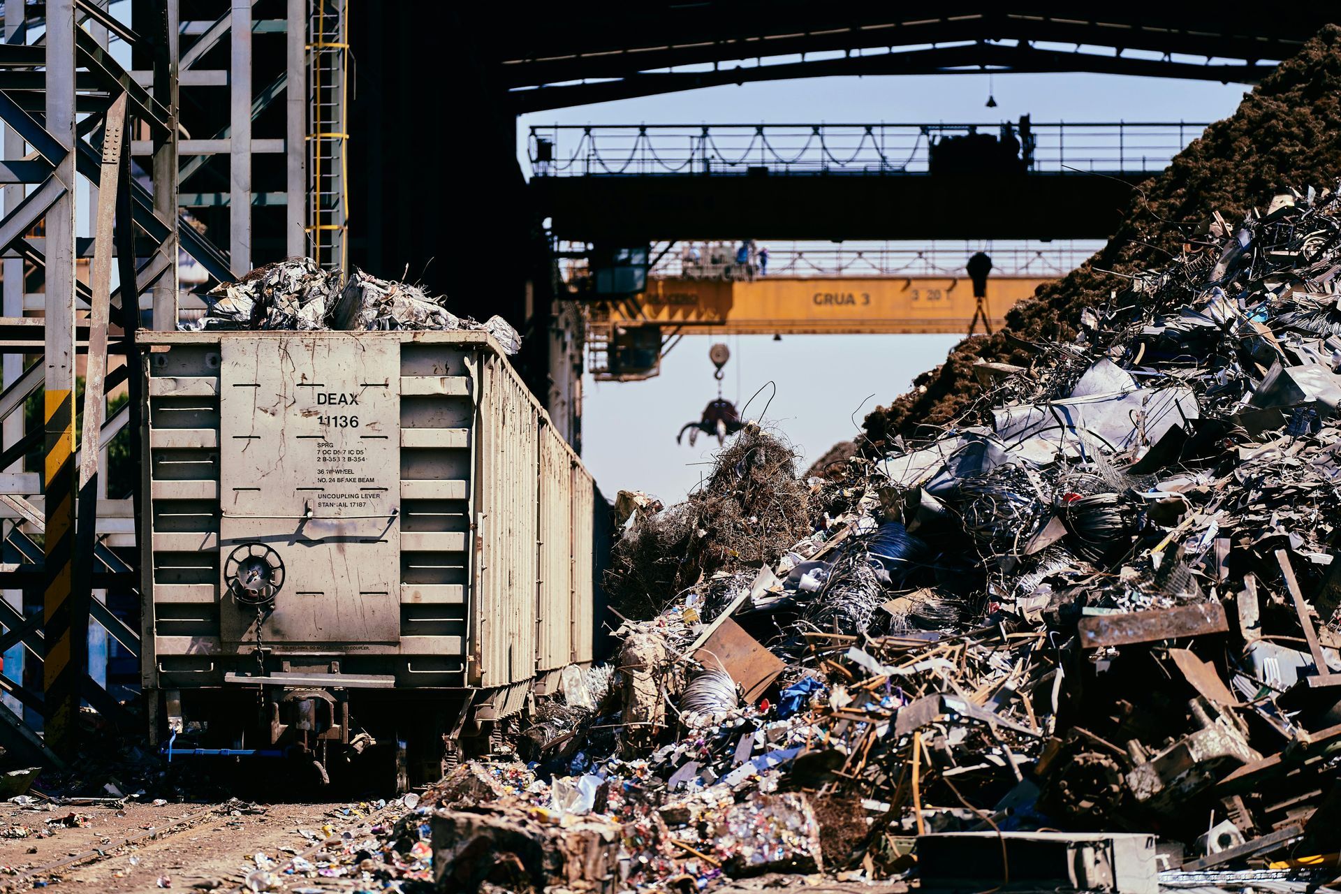 A large train car sits beside a massive pile of industrial scrap metal under a yellow overhead crane.