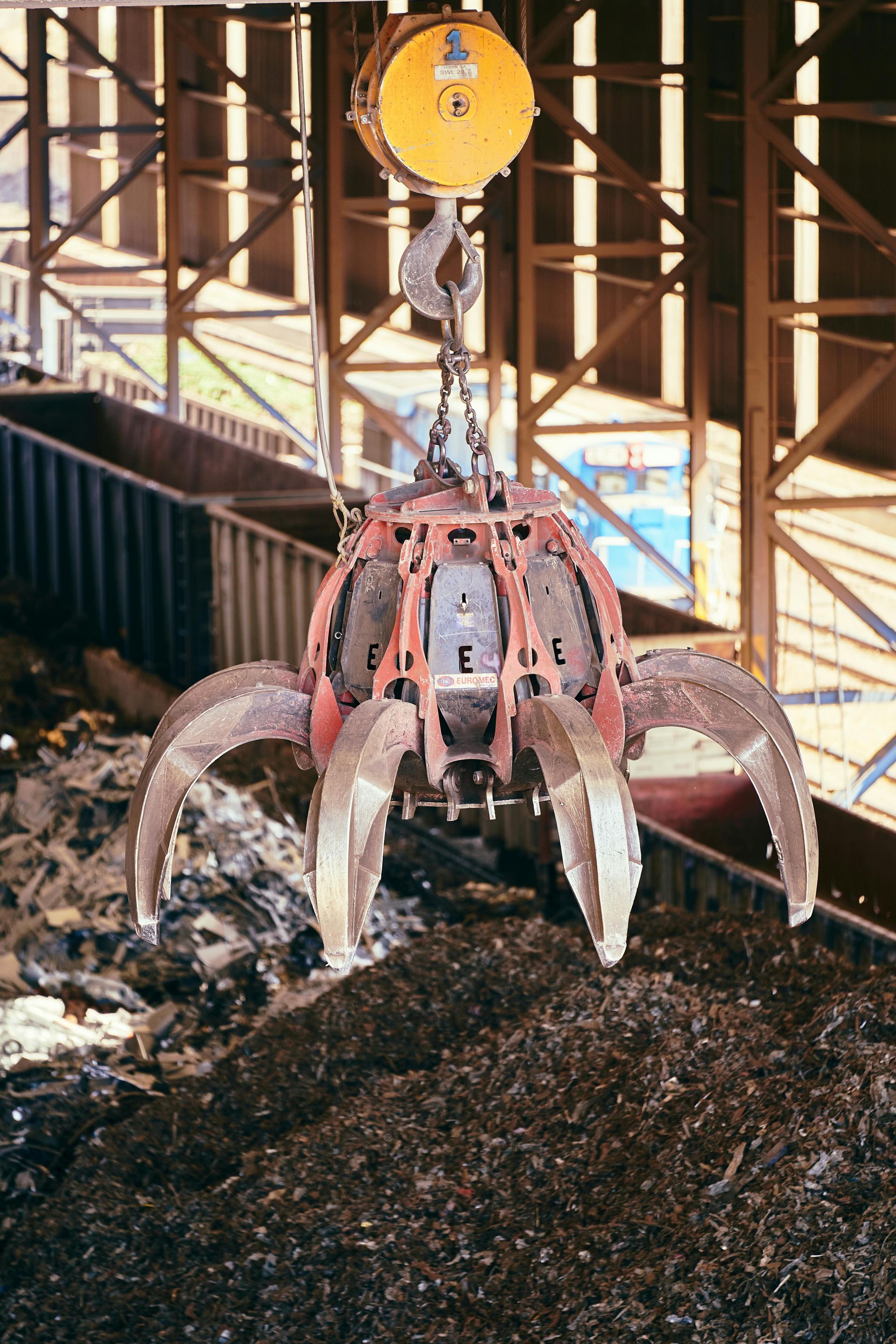 A yellow industrial orange-peel grab crane suspended over a pile of scrap metal in a warehouse.