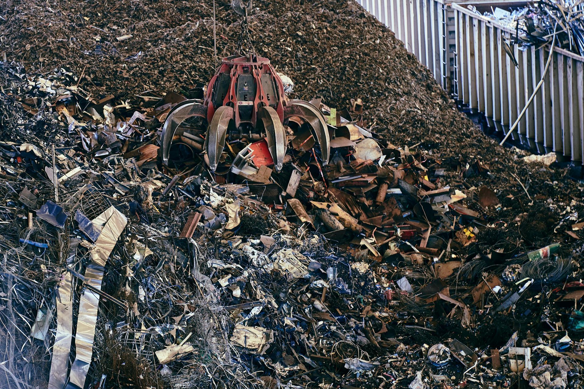 A large red industrial scrap grapple crane sorting through a massive pile of debris in a recycling yard.