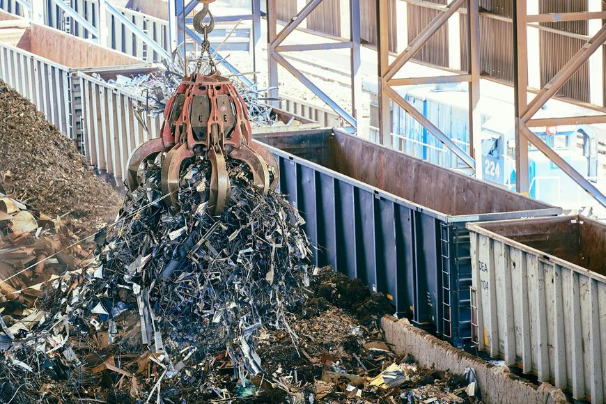 A large orange industrial grapple crane lifts a pile of scrap metal above railway cargo containers.