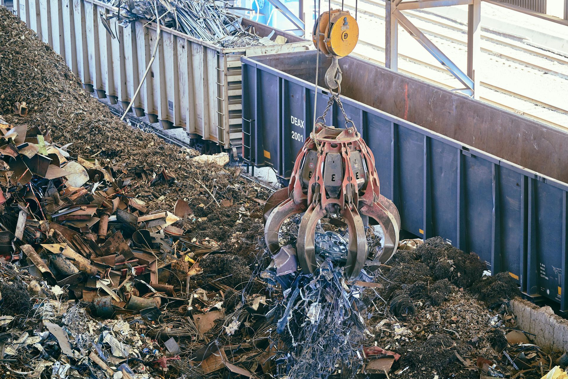 A mechanical claw suspended by a crane lifts scrap metal from a large pile into a train railcar.