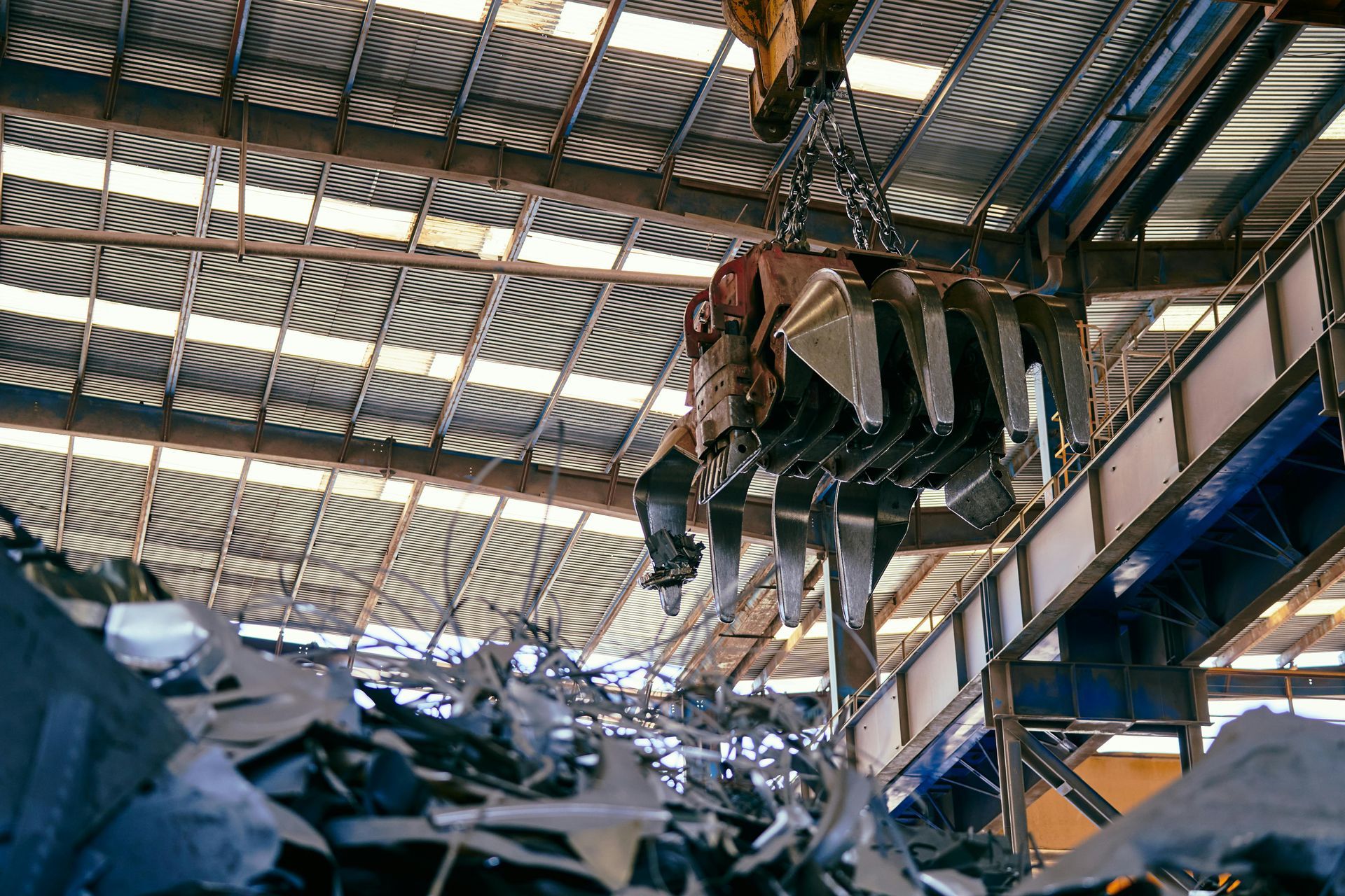 A large mechanical claw hangs from a ceiling crane in a warehouse, suspended over a massive pile of industrial scrap metal.