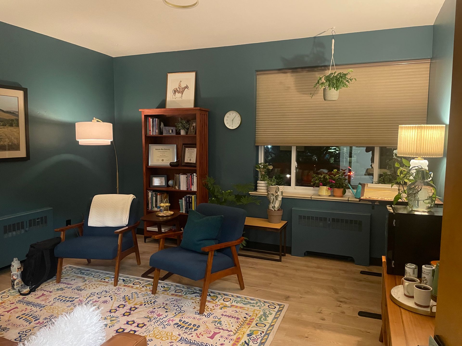 A cozy room with dark blue walls, two blue armchairs, a wooden bookcase, a colorful patterned rug, and warm lighting.