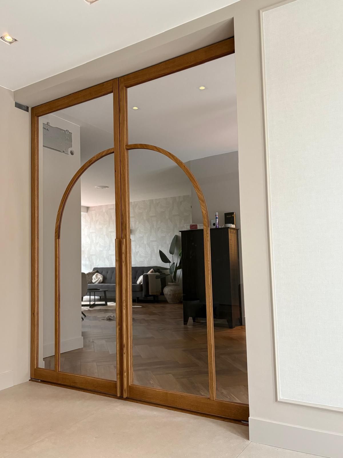 Glazen deuren met houten kozijnen en een boogvormige bovenzijde, die toegang geven tot een kamer met houten vloer en meubilair.