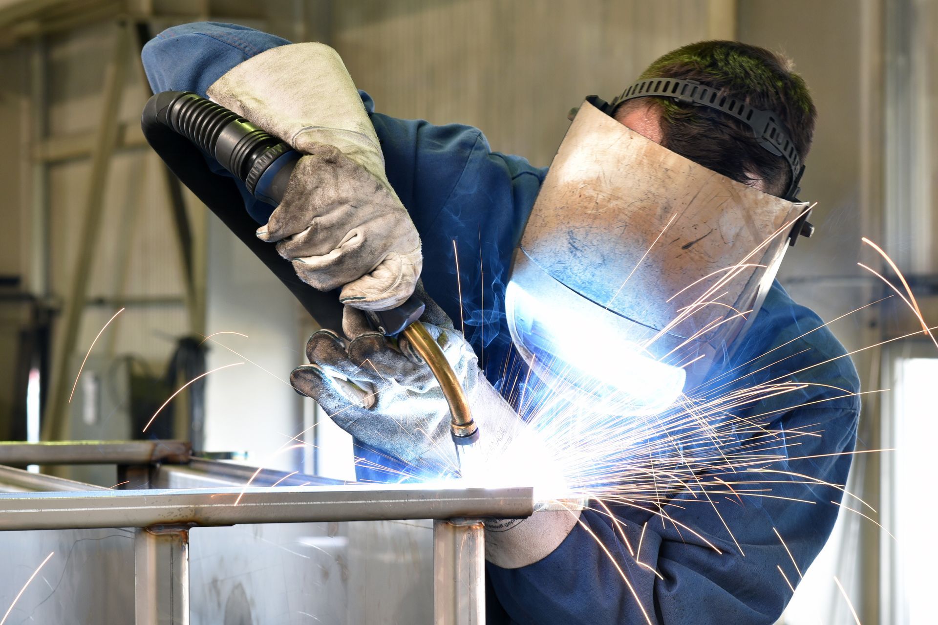 A welder in protective gear and a welding mask works on a metal frame, creating bright sparks in an industrial setting.