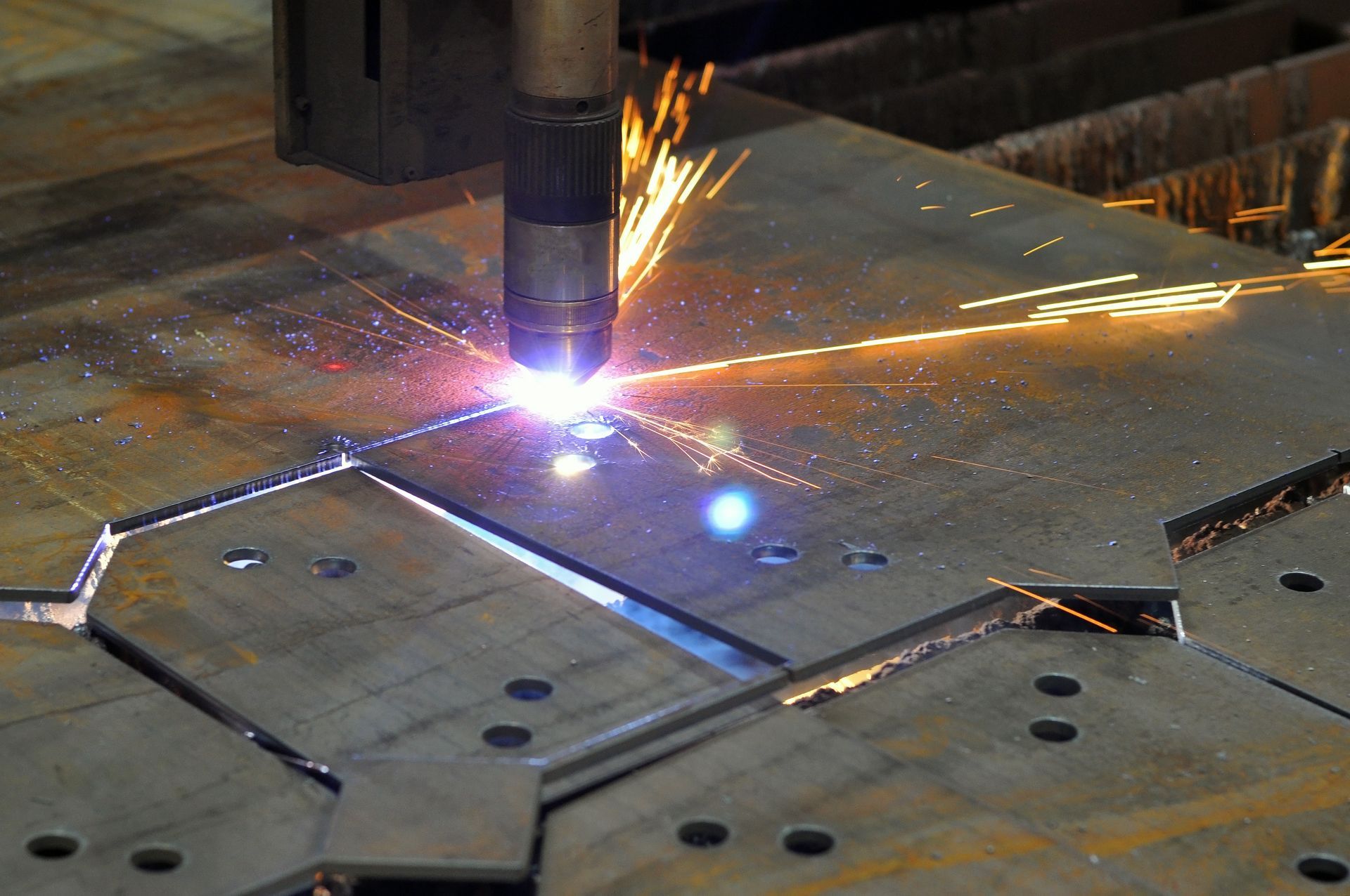 An industrial plasma cutter creates a glowing spark while precision-cutting shapes into a thick metal sheet.