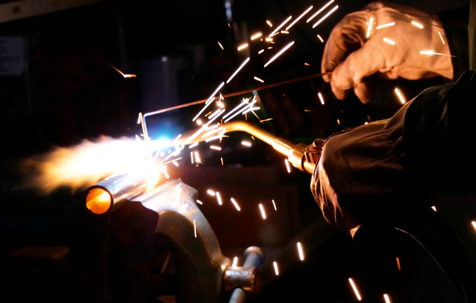 A gloved hand holds a torch to weld a metal pipe in a dark workshop, sending bright sparks flying into the air.
