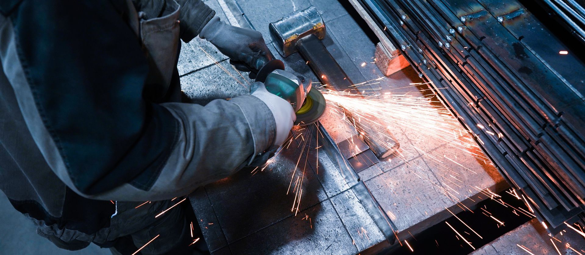 A person wearing protective gloves uses an angle grinder to cut a metal beam, producing a spray of bright orange sparks.