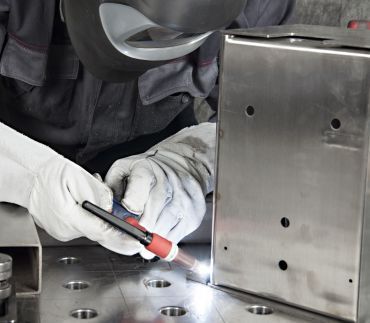 A welder in protective gear uses a TIG torch to weld a seam on a stainless steel metal box on a workshop table.