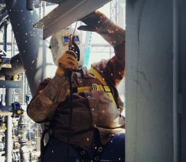 A welder in protective gear and a welding mask performs overhead welding on a steel structure in an industrial setting.