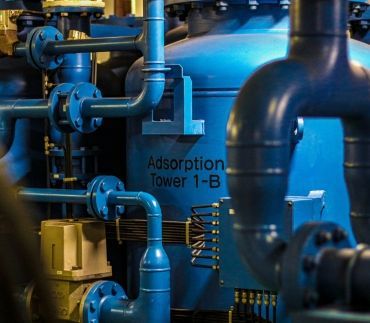A large, blue industrial adsorption tower with connected piping and wiring in a facility.