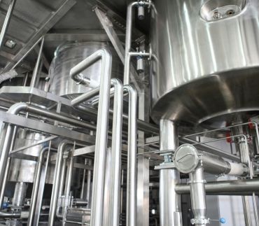 A low-angle view of industrial stainless steel tanks and complex piping systems in a manufacturing or brewing facility.