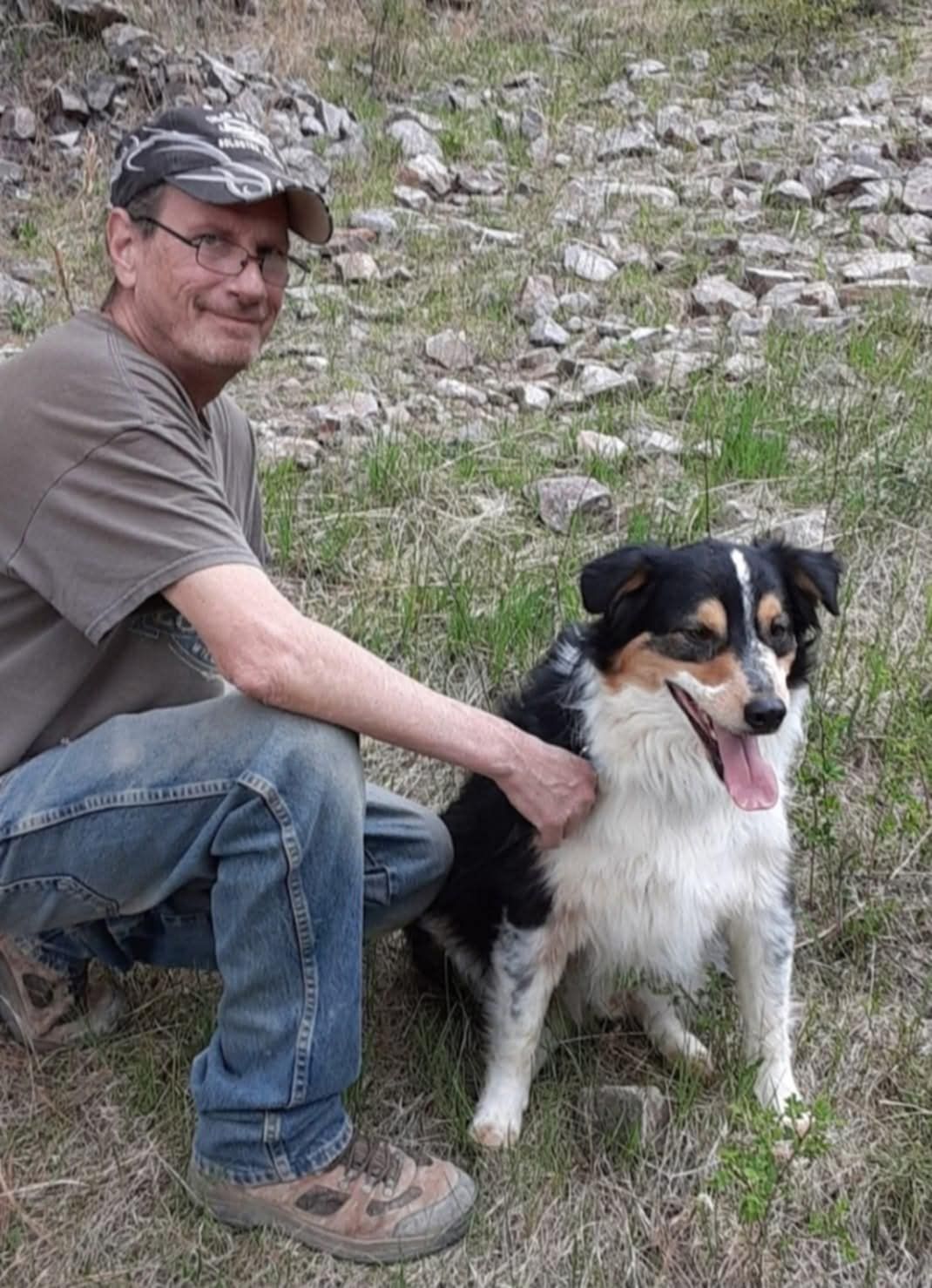 Man in cap and glasses kneels beside a tri-color dog outdoors on a rocky hillside. The man pets the smiling dog.