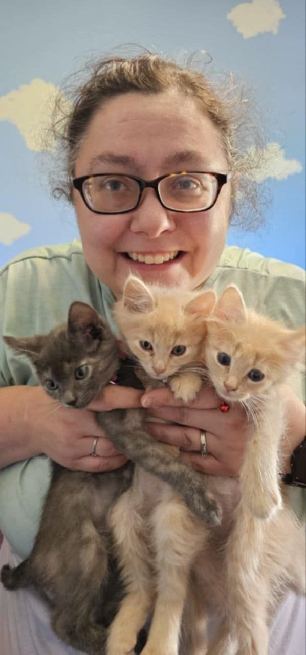 Woman with glasses smiles, a gray kitten sits on her shoulder, both in a car.