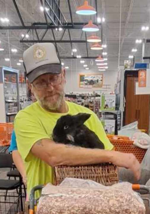 Man in a cap and yellow shirt holds a black rabbit in a basket at a store, with an orange shopping cart.