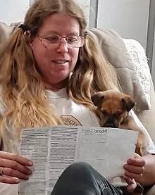 Woman with glasses reads paperwork, small brown dog rests on her shoulder.