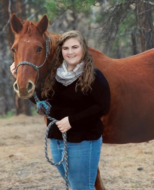 Woman in jeans and black sweater poses with a brown horse, holding its lead rope; outdoors.