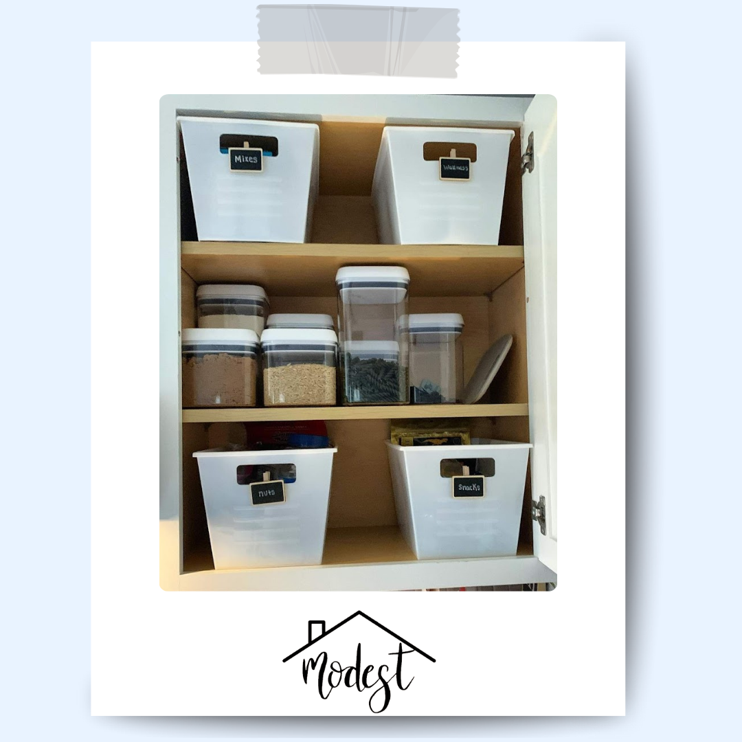 Organized kitchen cabinet with white containers and labeled jars on shelves.