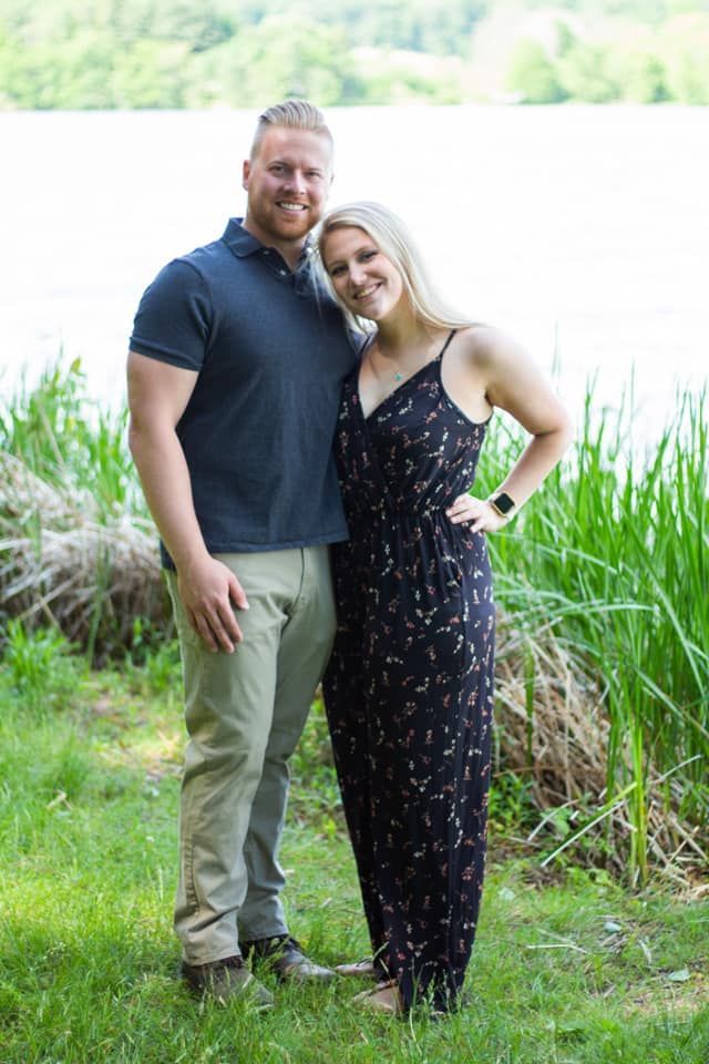 Man and woman smiling, posing by a lake. The man wears a navy shirt and khakis. The woman wears a black jumpsuit.