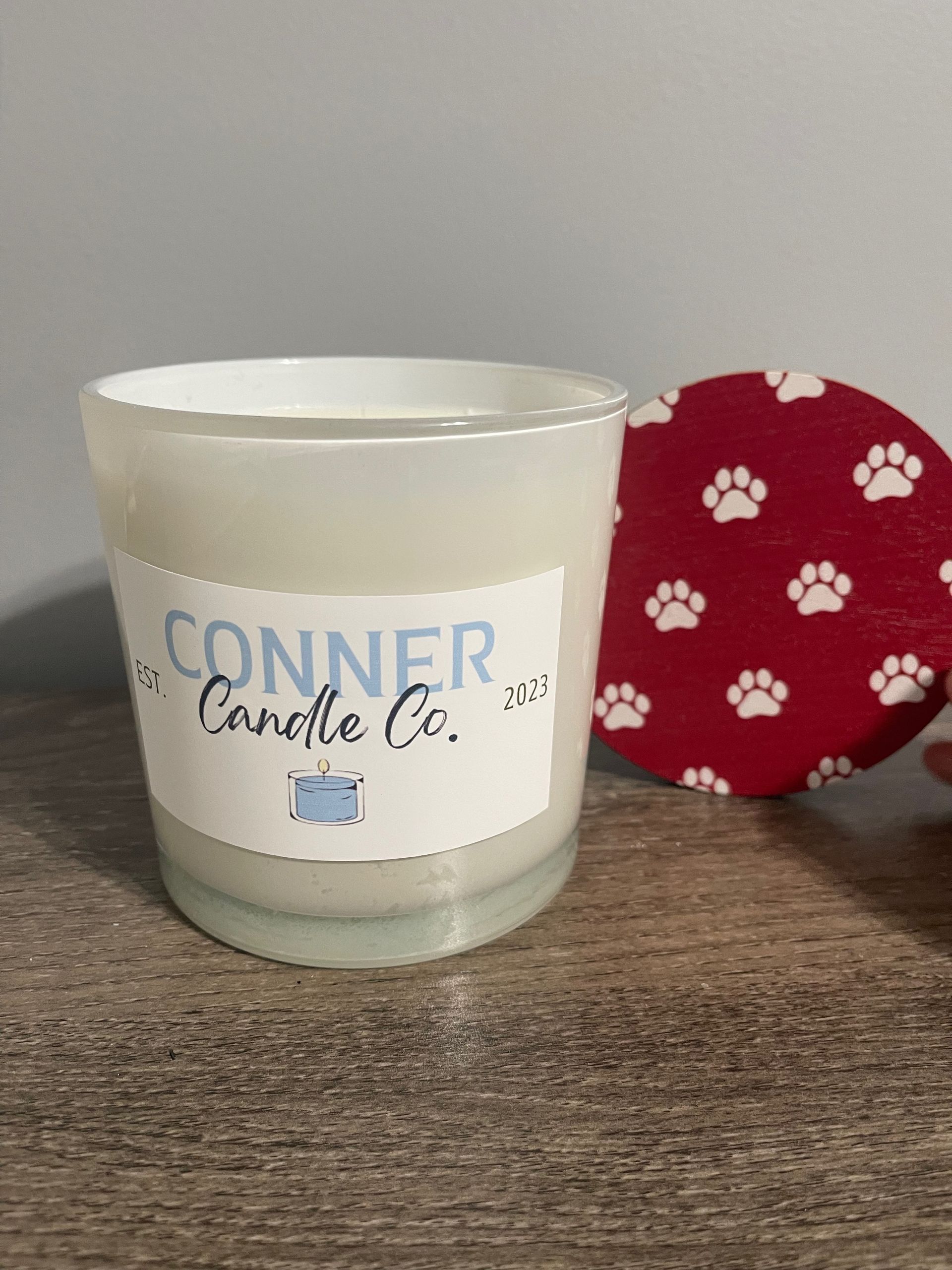 White candle with blue label, next to a red lid with white paw prints, on a wooden surface.